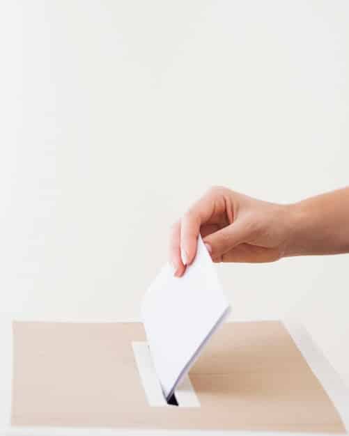 A close-up shot of a hand inserting a ballot into a voting machine. The focus is deliberately on the point of entry, emphasizing the physical act of voting. The background is intentionally blurred to keep focus on the hand and the machine. The lighting is neutral, and the atmosphere is functional and slightly sterile.