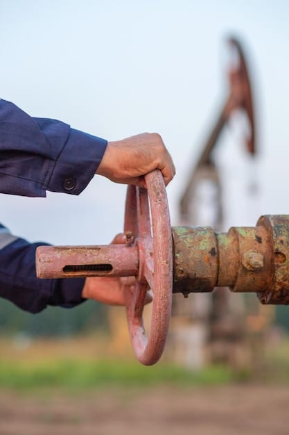 A close-up shot of a hand turning a valve on an oil pipeline, with a blurred background showing an industrial oil refinery.